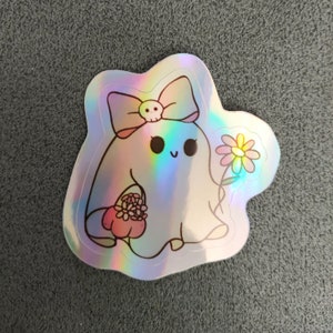 Puede incluir: Una pegatina hologr&aacute;fica con un lindo fantasma de dibujos animados que lleva un lazo rosa con un dije de calavera y sostiene un ramo de flores rosas. El fantasma tiene una expresi&oacute;n feliz y est&aacute; rodeado de un fondo iridiscente arco&iacute;ris.