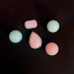 May include: An assortment of pastel-colored, small, smooth, and textured candies. The shapes include two spheres, a rectangle, a teardrop, and a round shape. The candies are light pink and light blue, set against a dark background.