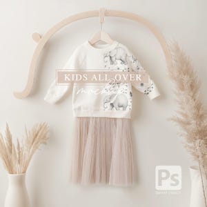 May include: A cream-coloured children's outfit comprising a long-sleeved sweatshirt and a skirt. The sweatshirt features a grey elephant graphic and the text "KIDS ALL OVER mockups". The skirt is light pink tulle. The outfit is suspended on a wooden hanger.