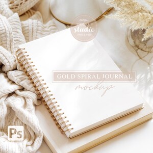 May include: A white spiral bound journal with gold rings, lying on top of a beige book. The journal is blank and ready for writing. The text "GOLD SPIRAL JOURNAL MOCKUP" is printed on the journal.