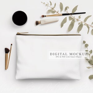 Könnte beinhalten: Weiße Canvas-Kosmetiktasche mit einem goldenen Reißverschluss. Die Tasche liegt flach auf einer weißen Oberfläche mit einem Zweig grüner Blätter und Make-up-Pinseln.