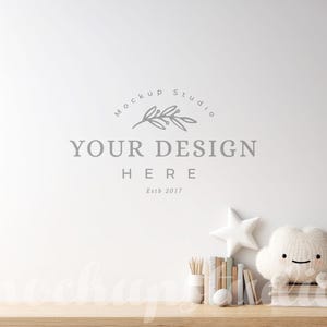 Könnte beinhalten: Eine Mockup-Studio-Szene mit einer weißen Wand mit dem Text "YOUR DESIGN HERE" und einem Logo. Ein Holzregal enthält ein wolkenförmiges Plüschtier, einen weißen Stern, Bücher und eine kleine Vase mit Bleistiften. Auf dem Regal befindet sich auch ein weißer Ball.