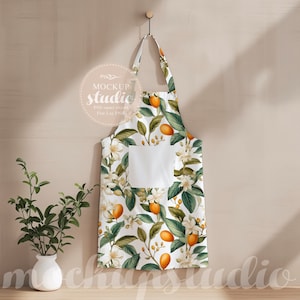 Op de afbeelding: Een witte schort met een bloemenpatroon met oranje citrusvruchten en witte bloemen. De schort heeft een grote zak aan de voorkant en hangt aan een haak tegen een lichtbeige muur.