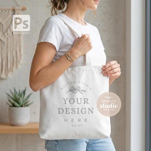 May include: A white tote bag with the text "YOUR DESIGN HERE" and a leaf design. The bag is held by a person wearing a white t-shirt. The bag has a Mockup Studio logo and the text "Estb 2017".