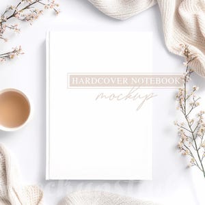 May include: White hardcover notebook mockup on a white surface.  The notebook is surrounded by beige knit fabric and small white flowers. Text on the notebook says 'HARDCOVER NOTEBOOK mockup' in a light brown serif font.  The overall aesthetic is minimalist and clean.