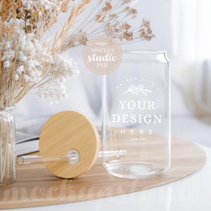 Puede incluir: Una lata de vidrio transparente con una tapa de bambú y un diseño blanco que dice "Your Design Here" y "Mock Up Studio Est. 2017". La lata está sentada en una bandeja de madera con flores secas en un jarrón en el fondo.