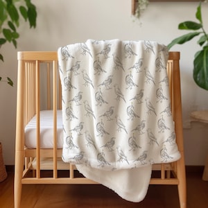 May include: A white baby blanket with a gray bird pattern. The blanket is folded and draped over a wooden crib.