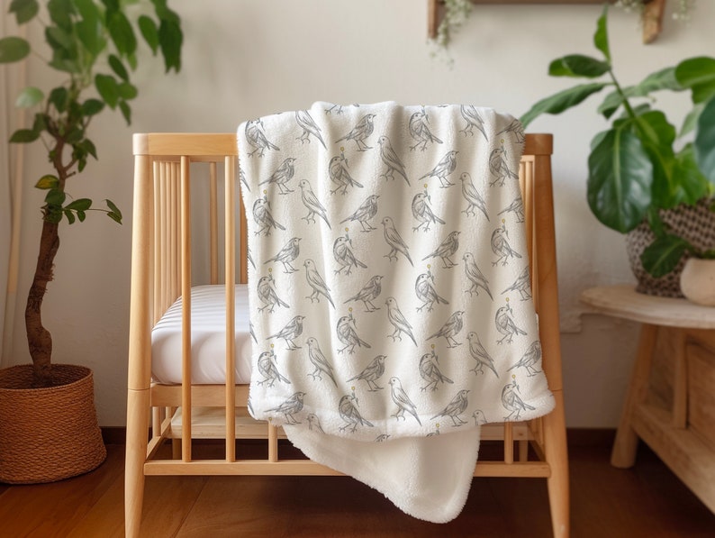 May include: A white baby blanket with a gray bird pattern. The blanket is folded and draped over a wooden crib.