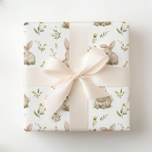 Peut inclure: Un cadeau emball&eacute; avec un ruban et un n&oelig;ud cr&egrave;me. Le papier d'emballage pr&eacute;sente un motif r&eacute;p&eacute;t&eacute; de lapins &agrave; l'aquarelle, de fleurs jaunes et de feuillage vert sur fond blanc. Le n&oelig;ud est centr&eacute; sur le cadeau.