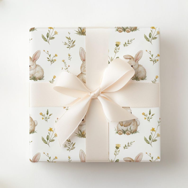 Peut inclure: Un cadeau emball&eacute; avec un ruban et un n&oelig;ud cr&egrave;me. Le papier d'emballage pr&eacute;sente un motif r&eacute;p&eacute;t&eacute; de lapins &agrave; l'aquarelle, de fleurs jaunes et de feuillage vert sur fond blanc. Le n&oelig;ud est centr&eacute; sur le cadeau.