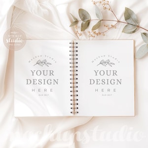 Puede incluir: Cuaderno abierto con espiral, ideal para maquetas de diseño. Presenta encuadernación dorada y páginas en blanco con el texto "YOUR DESIGN HERE". Descansa sobre tela blanca, con una ramita de follaje.
