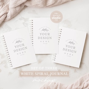 May include: Set of three white spiral bound journals with a simple design featuring the text "Your Design Here" and a small branch with leaves. The journals are on a white marble surface.