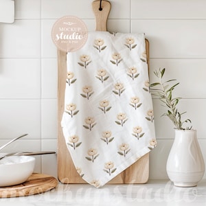 Puede incluir: Una toalla de cocina blanca con un patrón floral de flores beige sobre una tabla de cortar de madera. La toalla está colgando sobre el borde de la tabla.