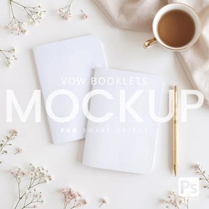 May include: A mockup featuring two blank white vow booklets, a gold pen, and a cup of tea with a gold rim. Delicate white and pink flowers and a beige cloth add to the aesthetic. The word "MOCKUP" is displayed in large, white letters.