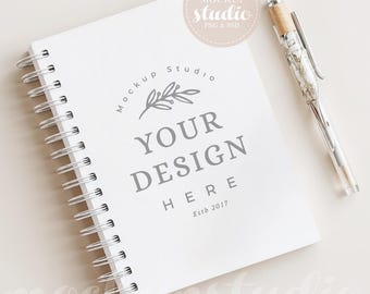 May include: A white spiral-bound notebook with the text "YOUR DESIGN HERE" and a silver pen with a marbled design. The notebook has a logo that says "Mockup Studio" and the text "Estb 2017".