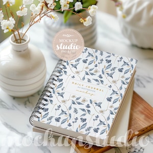 May include: A white spiral bound notebook with a blue and white floral pattern. The notebook has the text "Daily Journal A5 Notebook Mockup" on the front cover.