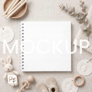May include: A square journal mockup with a white cover and spiral binding, surrounded by baby-themed items. Includes a plush bunny, wooden rattle, baby shoes, and tags with footprints. The text "SQUARE JOURNAL MOCKUP" is visible.