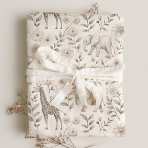 May include: A white muslin swaddle blanket with a brown and white floral pattern featuring giraffes and elephants. The blanket is tied with a white ribbon.