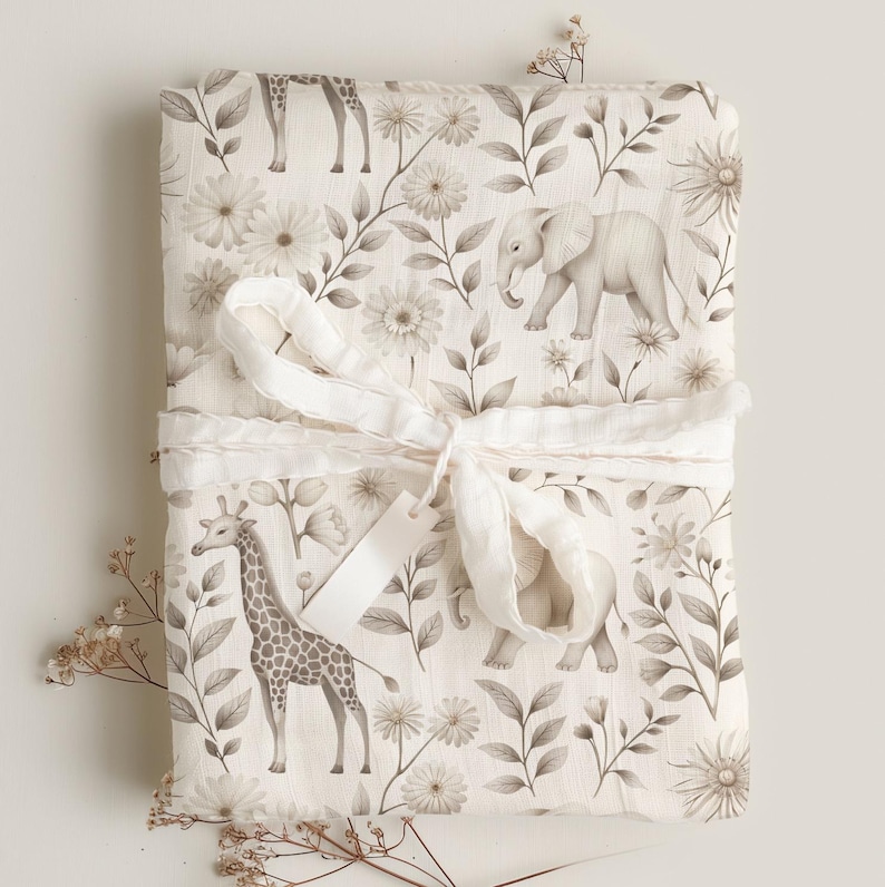 May include: A white muslin swaddle blanket with a brown and white floral pattern featuring giraffes and elephants. The blanket is tied with a white ribbon.
