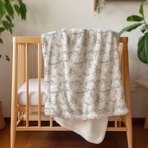 May include: A white and green floral patterned baby blanket with a soft white sherpa lining. The blanket is draped over a wooden crib.