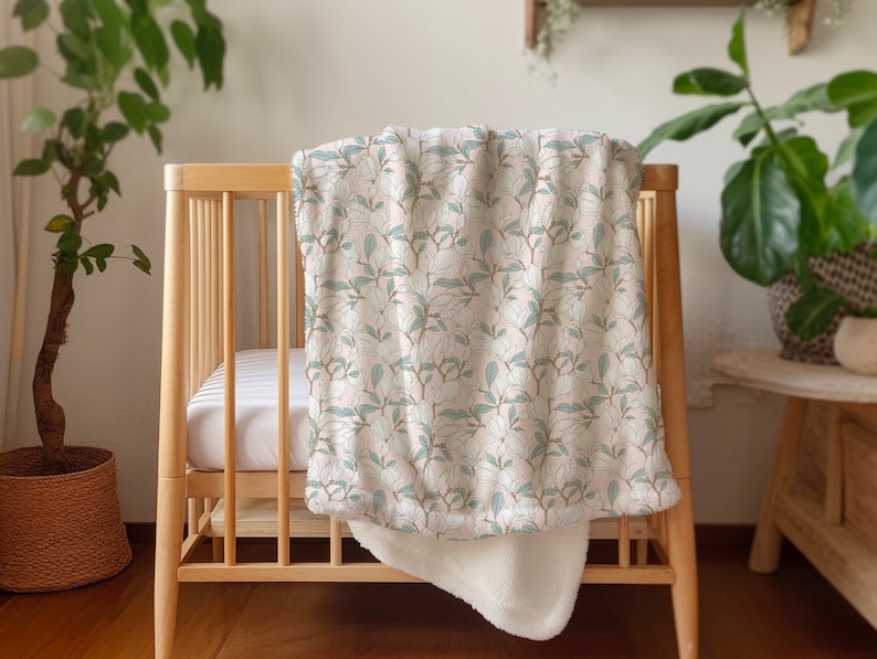 May include: A white and green floral patterned baby blanket with a soft white sherpa lining. The blanket is draped over a wooden crib.
