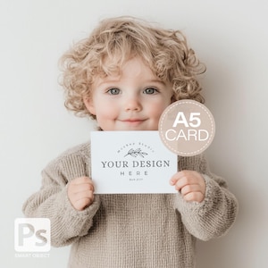May include: A child holding a white A5 card mockup. The card features the text "YOUR DESIGN HERE" and a floral design. The child is wearing a beige sweater and has curly blonde hair.
