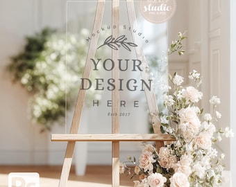 May include: A wooden easel holds a clear acrylic sign with the text "YOUR DESIGN HERE" and "Estb 2017". The sign is surrounded by white and pink flowers. A "Mockup Studio" logo is in the upper right corner.