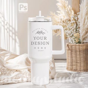 May include: White stainless steel travel mug with a handle and a straw. The mug has a white lid and a design that says "Mockup Studio" with a small branch and leaves, "Your Design Here", and "Est 2017".