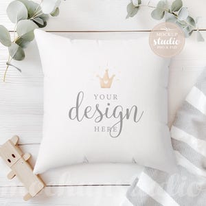 Op de afbeelding: Een wit kussen met een gouden kroon en de tekst "YOUR design HERE" op een wit houten oppervlak met groene eucalyptus takken en een wit-grijze gestreepte deken.