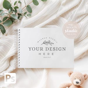 May include: A white spiral-bound notebook with the text "YOUR DESIGN HERE" and "Estb 2017". The notebook is on a white, textured surface with a sprig of green leaves and a small teddy bear. A "Mockup Studio" logo is visible.