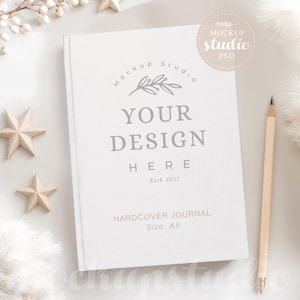 May include: A white hardcover journal mockup with the text "Mockup Studio", "Your Design Here", "Estb 2017", "Hardcover Journal", and "Size: A5". The journal is on a white surface with white feathers and gold stars.