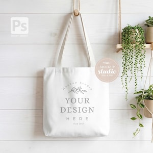 Może przedstawiać: Biała torba typu tote z napisem "YOUR DESIGN HERE" w szarej czcionce. Torba wisi na drewnianym kołku na białej ścianie z drewnianymi panelami. Projekt jest prosty i czysty, idealny do prezentacji niestandardowych grafik.