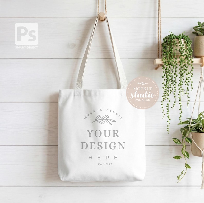 Może przedstawiać: Biała torba typu tote z napisem "YOUR DESIGN HERE" w szarej czcionce. Torba wisi na drewnianym kołku na białej ścianie z drewnianymi panelami. Projekt jest prosty i czysty, idealny do prezentacji niestandardowych grafik.
