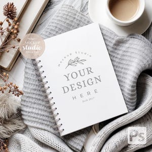 Può includere: Un taccuino bianco a spirale con il testo "Mockup Studio" e "Your Design Here" sulla copertina. Il taccuino è appoggiato su una coperta di maglia grigia con una tazza di caffè e un libro sullo sfondo.
