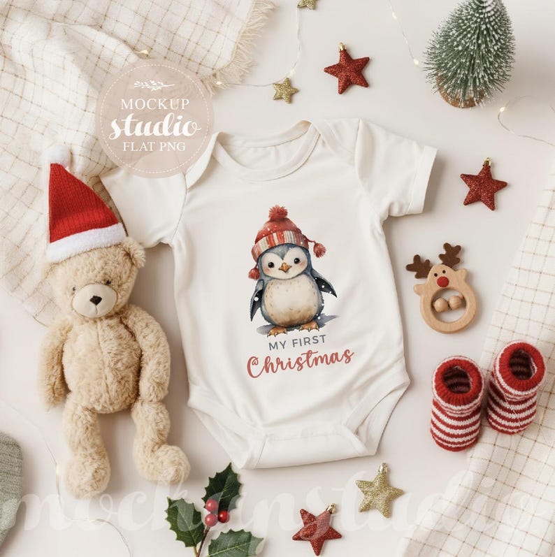 May include: A white baby onesie with a watercolor penguin wearing a red and white hat, with the text "My First Christmas." The image includes a teddy bear, Christmas decorations, and baby socks.