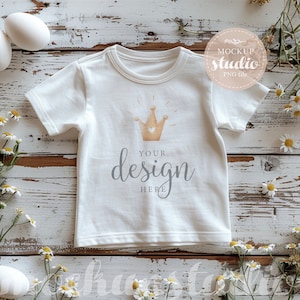 Könnte beinhalten: Weißes T-Shirt mit Rundhalsausschnitt, mit einem goldenen Kronen-Design und dem Text "your design here". Das Shirt ist von Gänseblümchen und Eiern auf einer weißen Holzoberfläche umgeben.