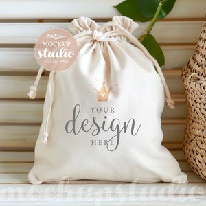 Puede incluir: Una bolsa blanca de cordón con una corona dorada y el texto "YOUR design HERE" impreso en ella. La bolsa está sobre una superficie de madera blanca.