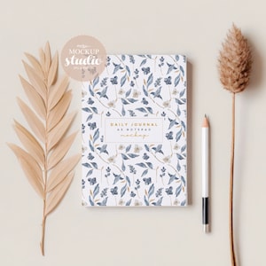 Puede incluir: Un diario con estampado floral con hojas azules y flores blancas, con la etiqueta "Daily Journal as Notepad". Un lápiz blanco con punta negra descansa junto al diario. También hay plantas secas y un logotipo de "Mockup Studio".
