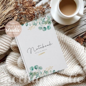 May include: A white notebook with a watercolor design of green leaves and gold accents. The notebook has the text "Notebook" and "PSD Digital Mockup" printed on the cover. The notebook is lying on a white knitted blanket with a cup of coffee and a plate on a wooden surface.