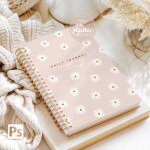 Puede incluir: Un diario de espiral rosa con un patrón de margaritas blancas y el texto "DAILY JOURNAL AS NOTEPAD MOCKUP" en la portada. El diario está sobre una superficie blanca con una manta de punto blanca y un jarrón con hierba seca.