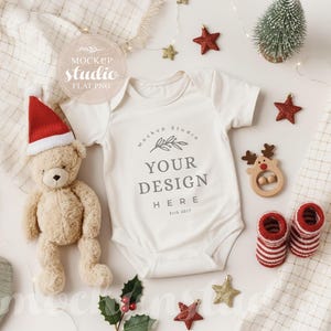 May include: A cream-colored baby onesie with the text "YOUR DESIGN HERE" is surrounded by Christmas-themed decorations. A teddy bear wearing a Santa hat, red star ornaments, and striped baby socks are visible.