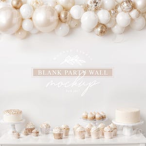 May include: A white wall with a garland of white and gold balloons. The wall has a logo that says "BLANK PARTY WALL MOCKUP". There is a table with a white cake and cupcakes with white frosting and gold sprinkles.