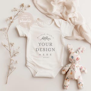 Pode incluir: Um macacão branco para bebê com mangas curtas e um design de maquete que diz "Mockup Studio, Your Design Here, Est. 2017". O macacão está colocado sobre uma superfície branca com um ramo de flores e um brinquedo de pelúcia de coelho.