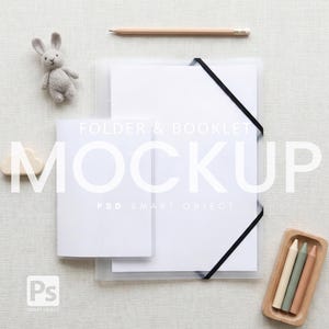May include: A mockup featuring a white folder and booklet with a black elastic band, a wooden pencil, a small gray knitted bunny, a wooden cloud, and a wooden tray with three crayons. The text "FOLDER & BOOKLET MOCKUP" is overlaid on the image.