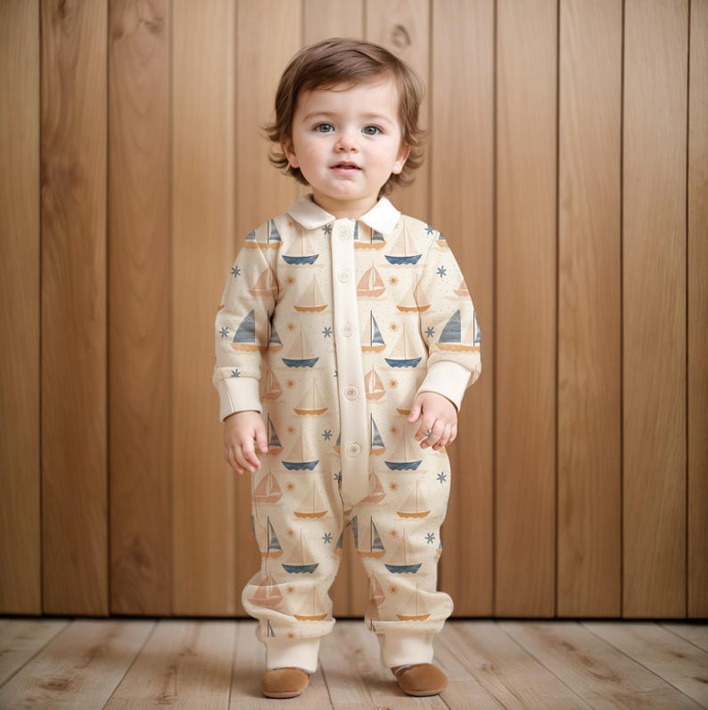 K&ouml;nnte beinhalten: Ein Baby tr&auml;gt einen cremefarbenen Strampler mit einem Muster aus Segelbooten und Sternen. Der Strampler hat einen wei&szlig;en Kragen und einen Knopfverschluss. Das Baby steht auf einem Holzboden.