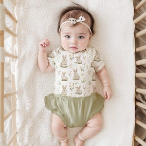Peut inclure: Un body pour b&eacute;b&eacute; avec un imprim&eacute; de lapins et de fleurs, associ&eacute; &agrave; une culotte &agrave; volants verte. Le b&eacute;b&eacute; porte un bandeau blanc avec un n&oelig;ud &agrave; pois noirs, couch&eacute; dans un lit b&eacute;b&eacute; en bois avec un drap &agrave; pois.