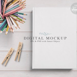 May include: A white book mockup with the text "DIGITAL MOCKUP JPG & PSD with Smart Object" on a white wood background. There are colorful pencils in a cup and wooden clothespins on the table.