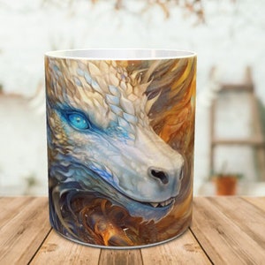 May include: White ceramic mug featuring a colourful illustration of a dragon's face. The dragon has blue eyes and scales, and its mouth is slightly open.
