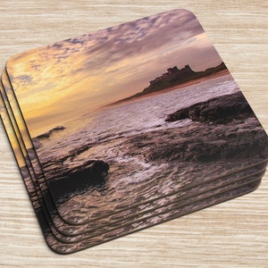 Könnte beinhalten: Ein Set mit vier quadratischen Untersetzern mit einem Foto einer Burg an einem felsigen Strand bei Sonnenuntergang. Der Himmel ist in leuchtenden Orange- und Rosatönen gehalten, das Wasser ist tiefblau. Die Burg ist vor dem Himmel silhouettiert.
