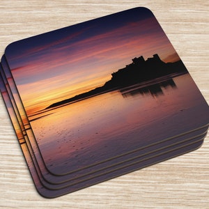 Könnte beinhalten: Ein Set mit vier quadratischen Untersetzern mit einem Foto einer Silhouette einer Burg an einem Strand bei Sonnenuntergang. Das Wasser spiegelt die Farben des Himmels wider, der eine Mischung aus Orange, Rosa und Lila ist.